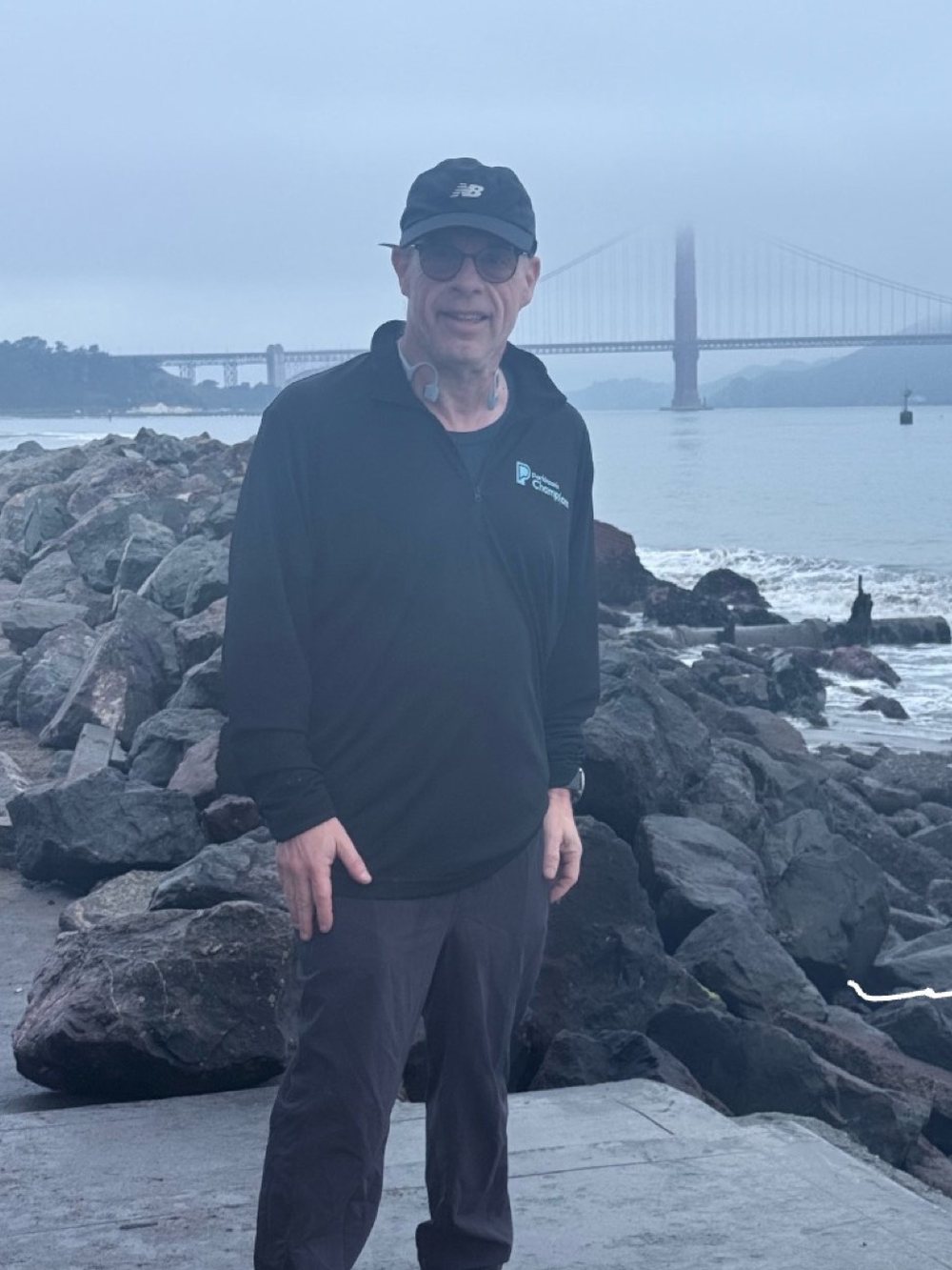 A runner in a Parkinson's Champions jacket standing by San Francisco Bay, the Golden Gate Bridge in fog behind him